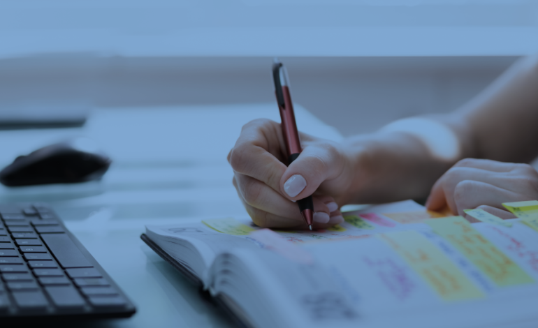 a person writes in a notebook with sticky notes, next to a computer keyboard